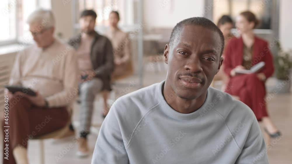 Chest-up of young male African corporate manager, looking on camera in foreground of blurred people sitting in conference room Chest-up of young male African corporate manager, looking on camera in foreground of blurred people sitting in conference room