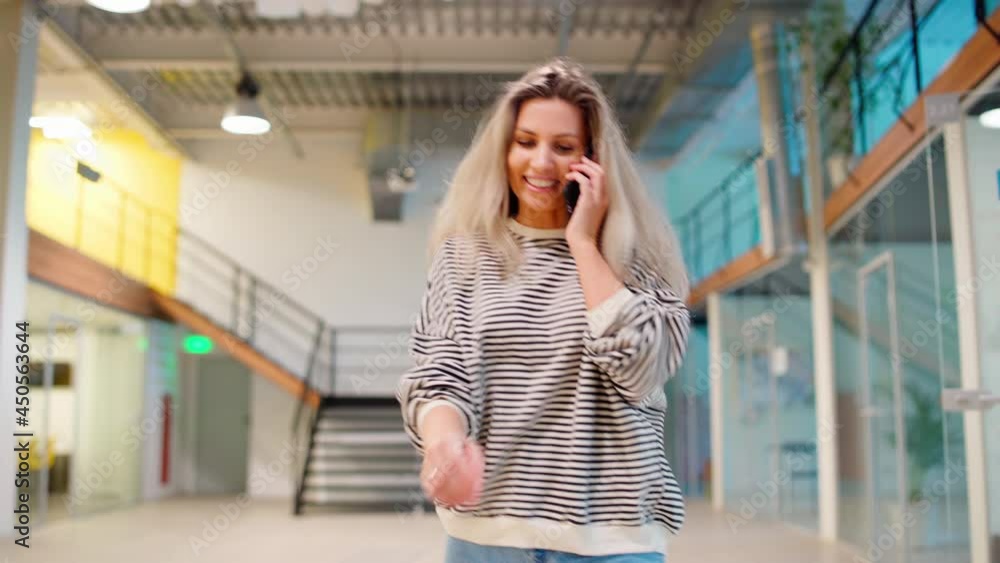 Blonde middle aged woman in striped sweatshirt walking down hall in modern office building and answering call on cell phone. Smiling female entrepreneur talking on phone at break Blonde middle aged woman in striped sweatshirt walking down hall in modern office building and answering call on cell phone. Smiling female entrepreneur talking on phone at break