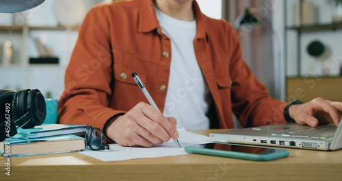 Unrecognizable diligent male student sitting near the computer and making needed notes into copybook while studing alone at home photo
