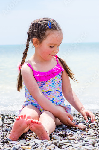 Girl 5 years old, barefoot, in bathing suit with pink ruffles with dark hair and plaited braids, hair clips and rubber bands on your hair playing on the shore of the sea with small pebbles in calm sun