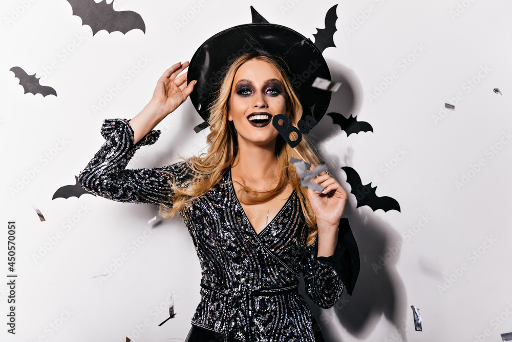 Excited glamorous witch with black makeup laughing to camera. Smiling blonde vampire in hat relaxing in halloween. Excited glamorous witch with black makeup laughing to camera. Smiling blonde vampire in hat relaxing in halloween.