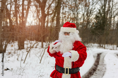 Santa Claus standing near a forest tree holding in a red bag gifts for children for Christmas around snow