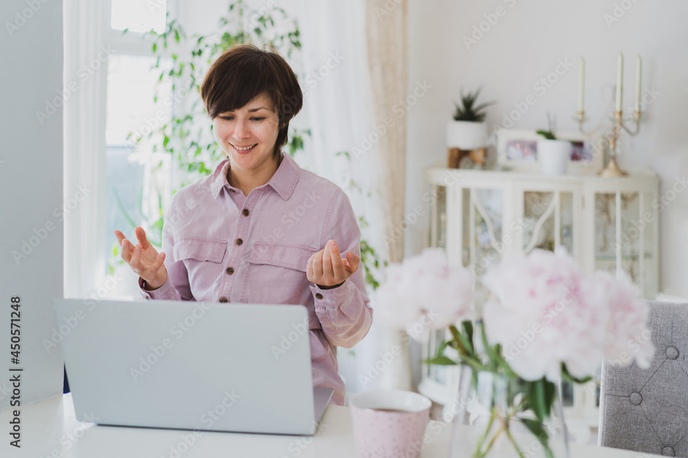 Businesswoman working at home, using laptop, gesturing, participating in an online meeting.. Cozy workspace with fresh peony flowers on the white table with light interior background. Remote work. Businesswoman working at home, using laptop, gesturing, participating in an online meeting.. Cozy workspace with fresh peony flowers on the white table with light interior background. Remote work.