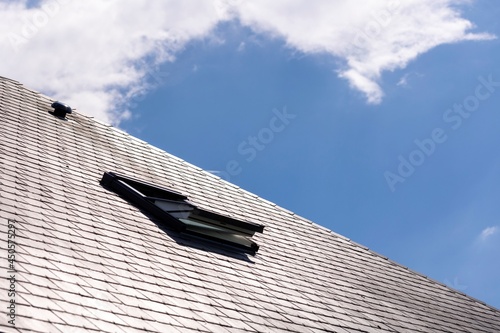 A portrait of an opened skylight window for ventilation in a slate roof on a sunny day with a blue sky with white clouds. you can also see a ventilation vent on the rooftop.