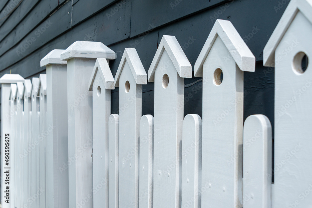 Fototapeta premium A white wooden fence with two gate posts in the center. The top of the fence has a small decorative birdhouse with a hole on the top of each paling. There's a navy blue house in the background.