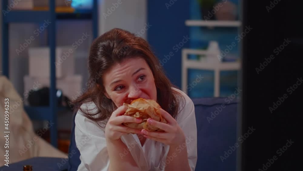 Portrait of young woman eating burger and drinking beer from bottle sitting on living room couch. Adult enjoying takeaway meal with alcoholic beverage while watching television after work Portrait of young woman eating burger and drinking beer from bottle sitting on living room couch. Adult enjoying takeaway meal with alcoholic beverage while watching television after work