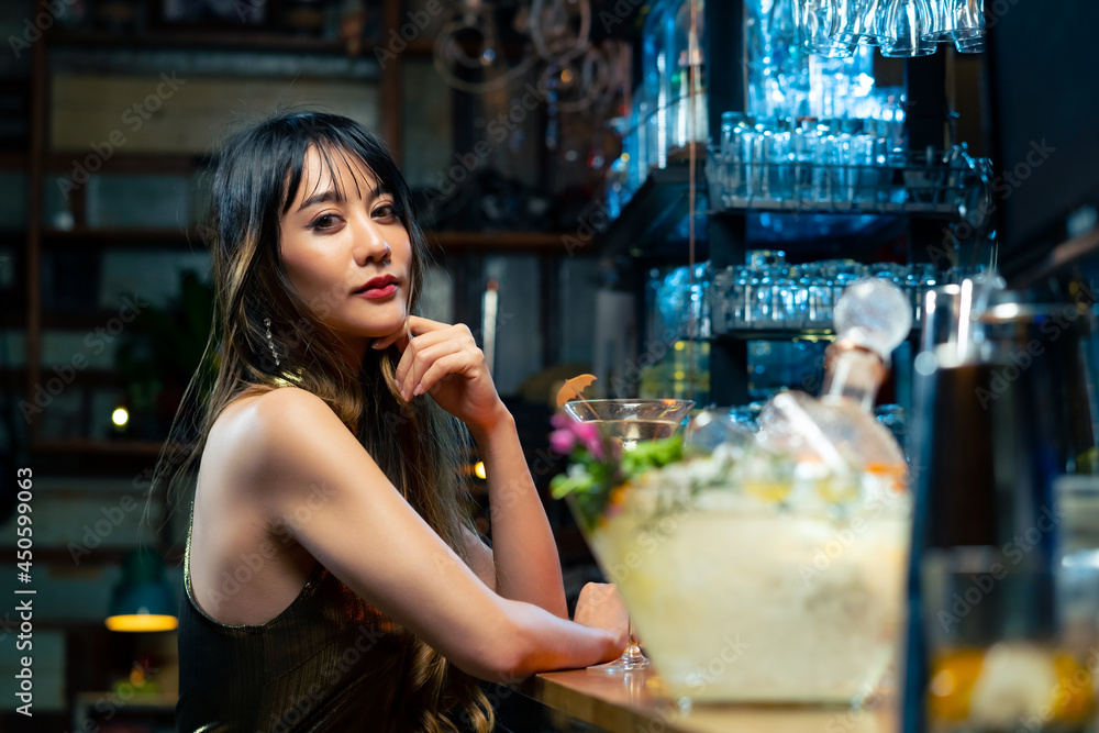 Asian woman sitting at bar counter drinking cocktail and talking to barman in nightclub. Male mixologist bartender preparing alcoholic drink to customer. Small business bar and city nightlife concept