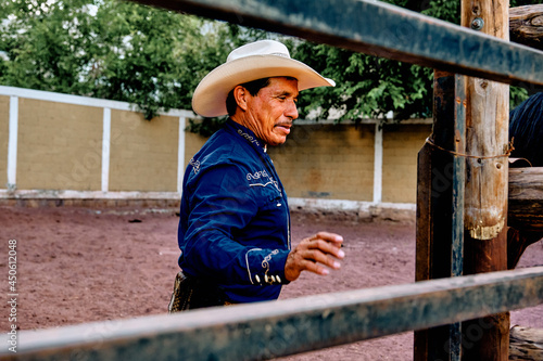 Mexican, hispanic, latino middle aged rancher in a blue shirt wearing a cowboy hat in a riding hall opening the fence for the horses