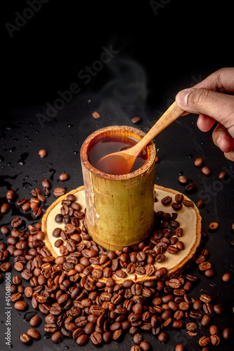 Hand holding wooden spoon and brew coffee, Many roasted coffee bean scattered on black background, dark coffee has smoke and aroma, Drink for break, coffee mugs mugs made from bamboo, local style.