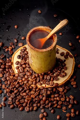Coffee mugs made from bamboo on wooden saucer and many roasted coffee bean scattered on black background, dark coffee has smoke and aroma, Drink for break, morning and coffee time, local style.