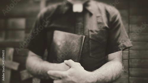Clergyman, reverend or priest wearing a clerical collar and clutching a bible. Preacher preaching the gospel in front of an old rustic rural church or cemetery.