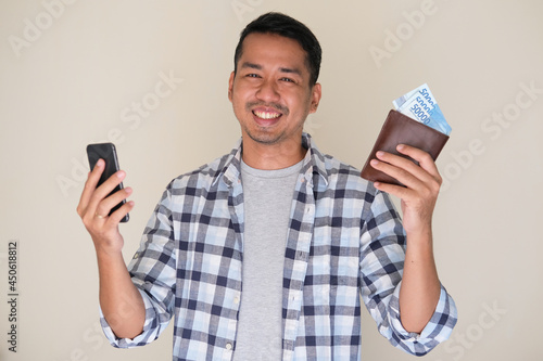 Adult Asian man smiling happy while holding mobile phone and showing paper money from his wallet photo