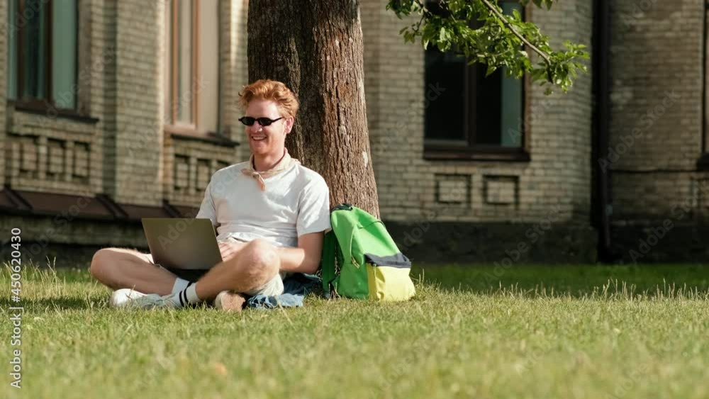 A happy man student is using his laptop sitting on the grass on campus outside A happy man student is using his laptop sitting on the grass on campus outside