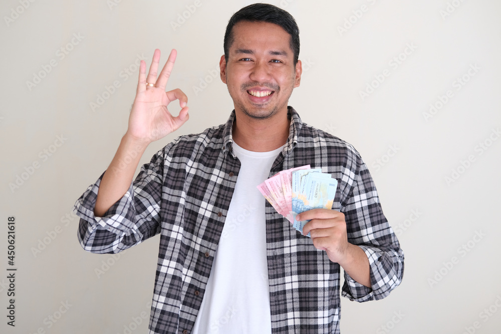 Adult asian man smiling happy giving “OK” sign while holding paper money Adult asian man smiling happy giving “OK” sign while holding paper money