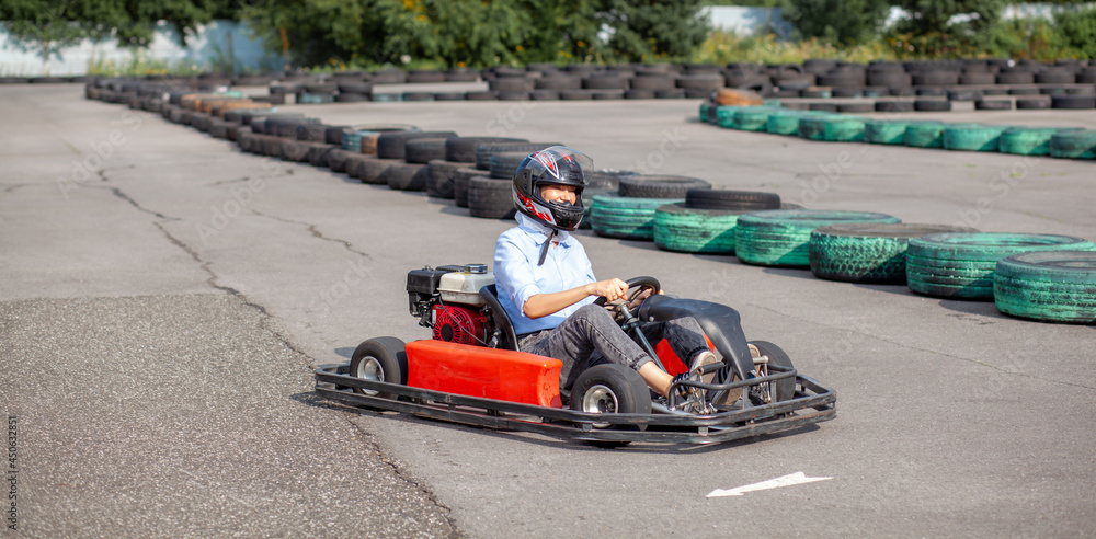 Fototapeta premium A girl or a woman in a hard hat rides a go-kart on a special track fenced with rubber wheels. Active recreation and sports on transport. Preparation and training for competitions.