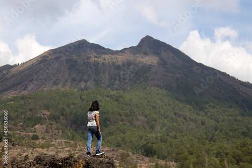 Woman standing alone on a rock looking at the blue mountains nature scenery. Happiness, strength and freedom lifestyle inspiraitonal concept. Lost in Mount Batur, Kintamani, Bali, Indonesia photo