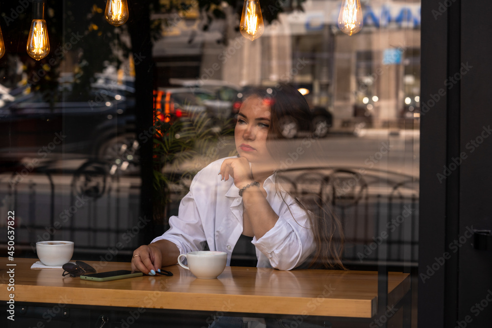 Woman at trendy cafe drinking coffee or tea. Young beautiful Caucasian model in white shirt and red lipstick. Reflections from windows. Image cross processed. Shot through the window of fashion girl. Woman at trendy cafe drinking coffee or tea. Young beautiful Caucasian model in white shirt and red lipstick. Reflections from windows. Image cross processed. Shot through the window of fashion girl.
