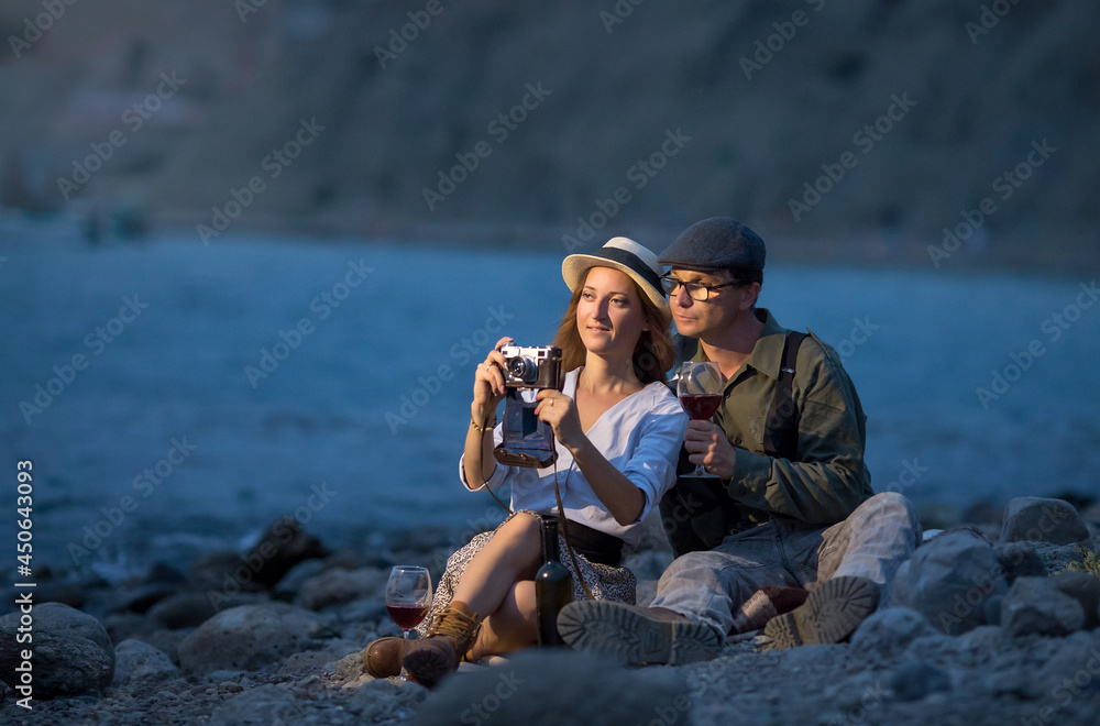 a young man and a woman drink red wine on the beach from glasses, sitting on a blanket in the evening, they have a camera, take pictures of the landscape.