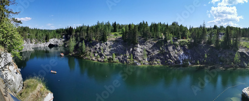 Panorama with a view of the rocks, grotto and turquoise water of the Marble Canyon in the Ruskeala Mountain Park, where boats float and the sky and trees are reflected on a sunny summer day.