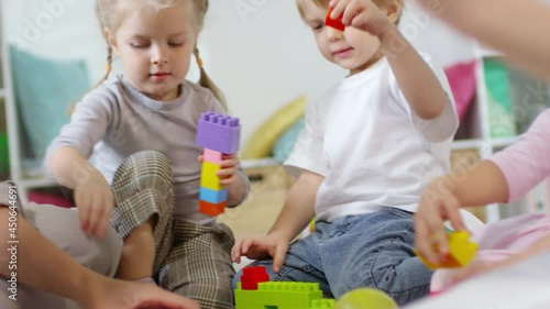 Close-up view of cute little boys and girls sitting on the floor in kindergarten and building something with colorful plastic bricks while playing developing game with help of female teacher photo