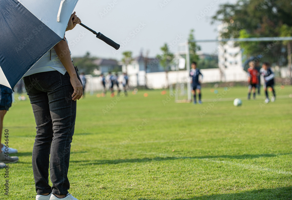Obraz premium Mother standing and watching her son playing football in a school tournament on a clear sky and sunny day. Sport, active lifestyle, happy family and soccer mom concept.