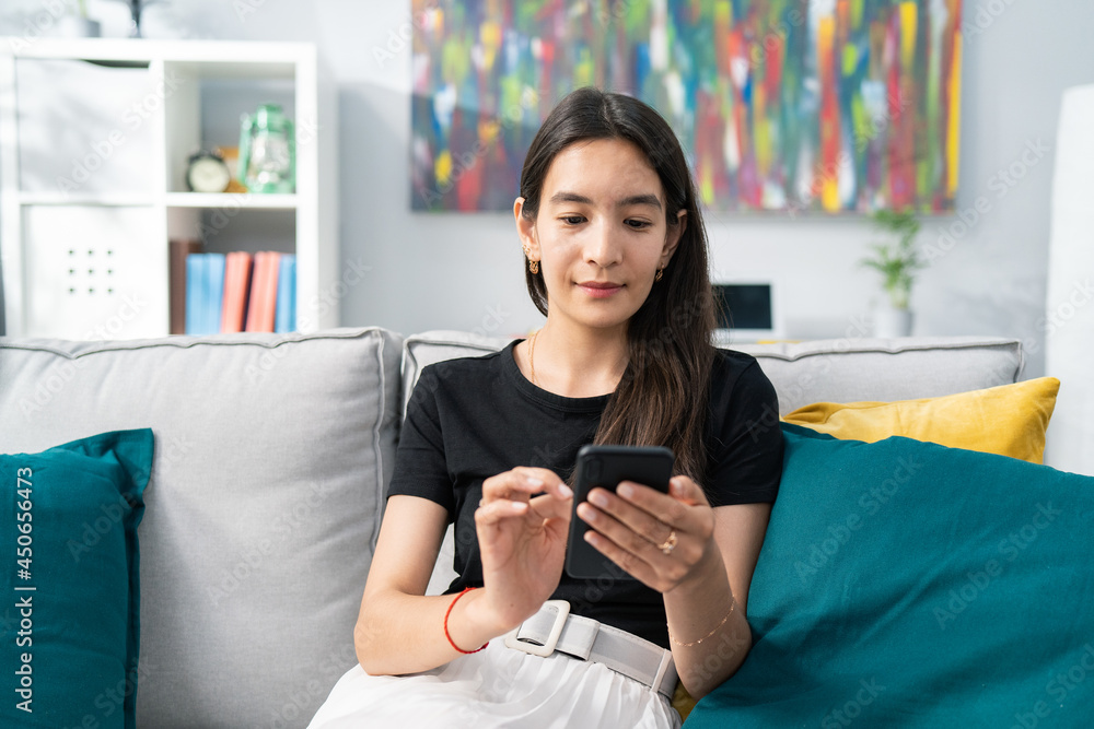 A beautiful Asian woman with brunette hair sits smiling gently on a couch in living room, dressed in linen skirt and loose black tshirt, holding phone in hands, texting with friends, chatting A beautiful Asian woman with brunette hair sits smiling gently on a couch in living room, dressed in linen skirt and loose black tshirt, holding phone in hands, texting with friends, chatting