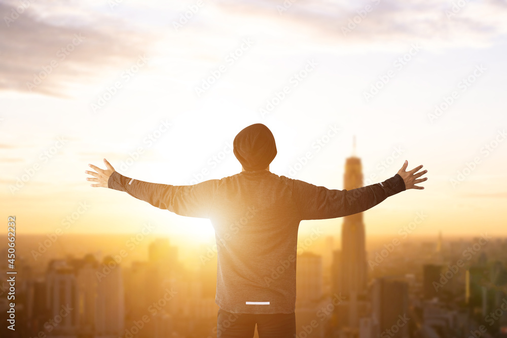 Silhouette of young businessman wearing sweater raise both hands and watched beautiful view sunset over office building. He likes to stand at the top of the building and enjoy the view.