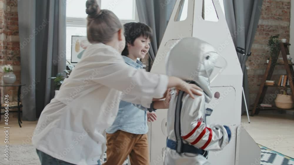 Handheld slowmo of group of happy kids running around cardboard rocket ship standing in center of living room. One boy is wearing spacesuit