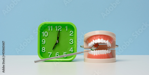 green alarm clock and a plastic model of a human jaw with white even teeth and a medical examination mirror on a white table. Morning teeth cleaning