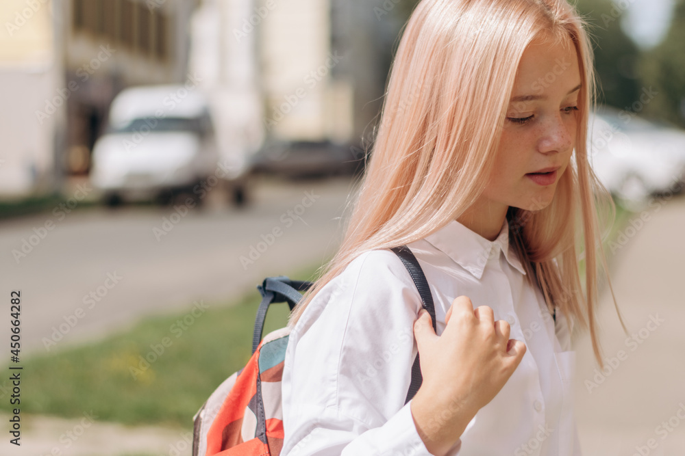 Fototapeta premium Serious blonde teenage girl in a school uniform and a backpack is walking down the street to school.Back to school concept.Copy space for text.