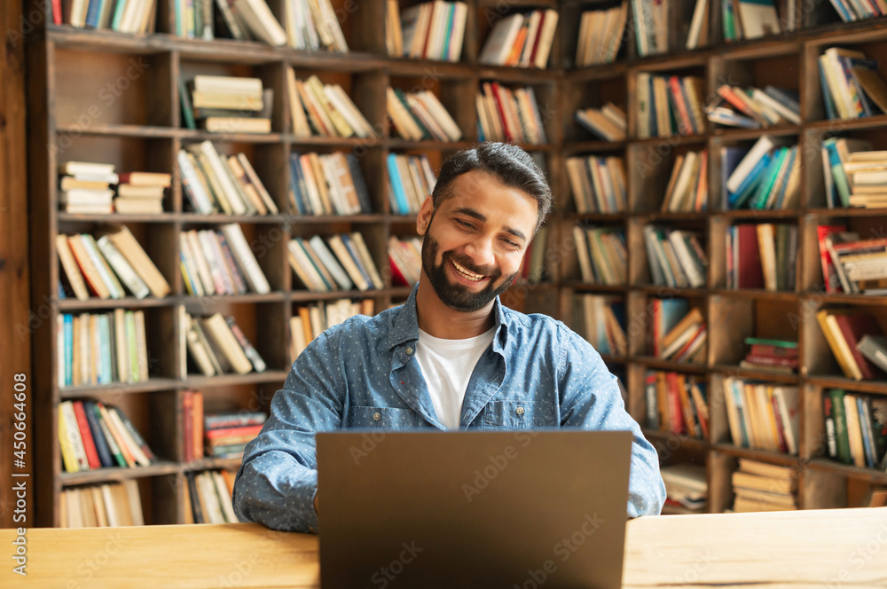 Cheerful Indian male freelancer looks at the laptop screen, eastern student websurfing sitting at the desk in the library, arabic guy looking through job offers vacancies, bookshelves on background Cheerful Indian male freelancer looks at the laptop screen, eastern student websurfing sitting at the desk in the library, arabic guy looking through job offers vacancies, bookshelves on background