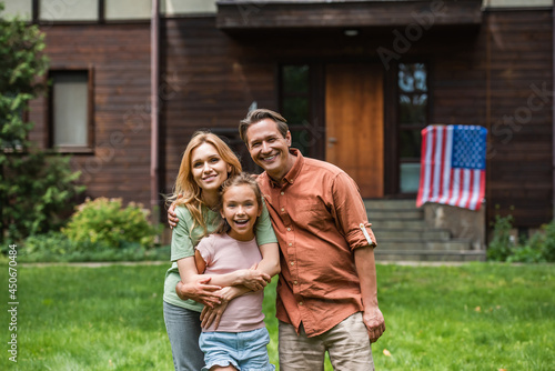 Smiling family hugging near blurred american flag on house outdoors