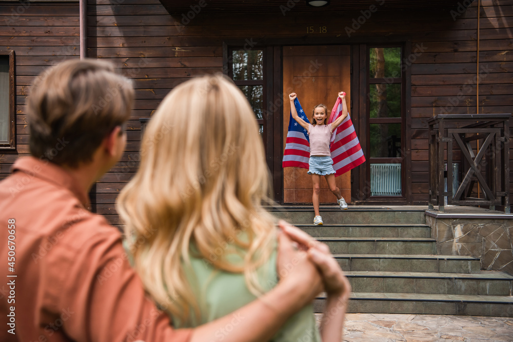 Excited girl holding american flag near blurred parents embracing outdoors