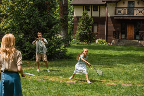 Kid and mother playing badminton near father with smartphone outdoors