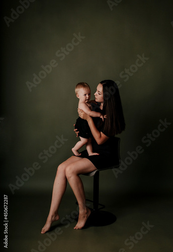 a brunette mom in a black dress is sitting with her baby daughter on a green background with a place for text