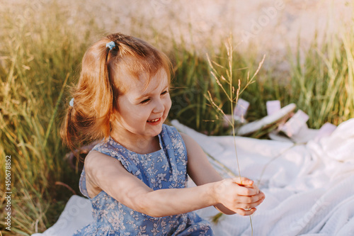 Little girl enjoy summer in field with wild green grass. Caucasian baby wear blue eco cotton dress holding blade and smiling
