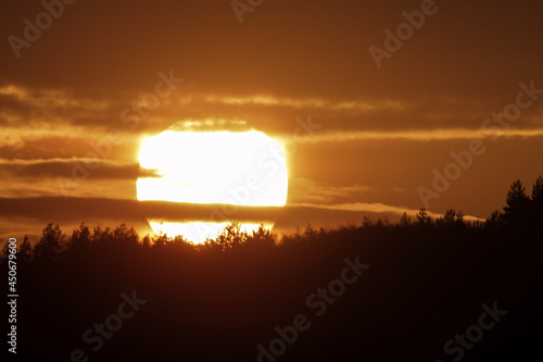 A large solar disk sets behind the mountains and the forest on the horizon. Red sunset sky.