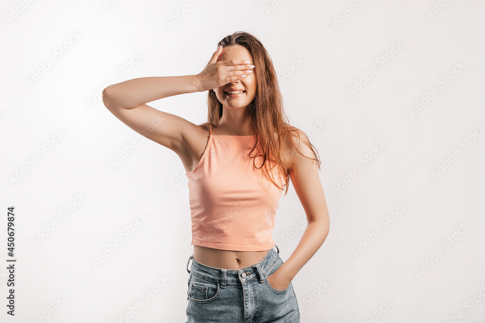 Young beautiful girl smiles and covers of her face with her hand palm on a white isolated background. Positive brunette woman in orange top Young beautiful girl smiles and covers of her face with her hand palm on a white isolated background. Positive brunette woman in orange top