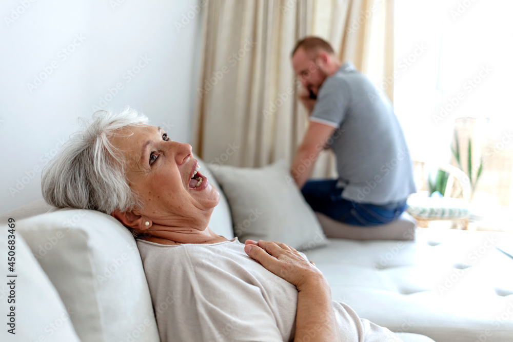 Obraz premium Shocked young man holding phone calling emergency ambulance worried about his grandma at home during the day. Young man asking for help from a sick senior woman. Healthcare and medicine concept.