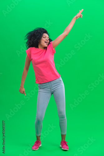 Portrait of young female African American cheerfully dancing the dance of victory, joy or good mood. Black woman with curly hair poses on green screen in studio. Full length.