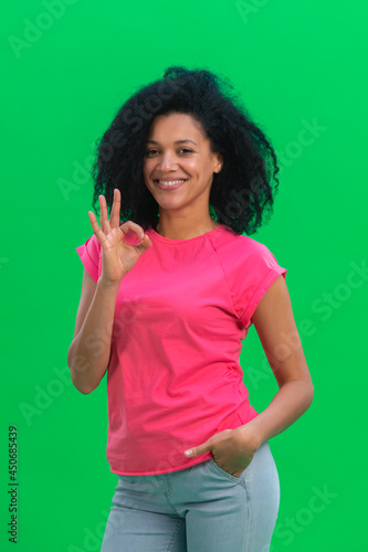 Portrait of young female African American smiling and making sign ok. Black woman with curly hair in pink tshirt poses on green screen in the studio. Close up.