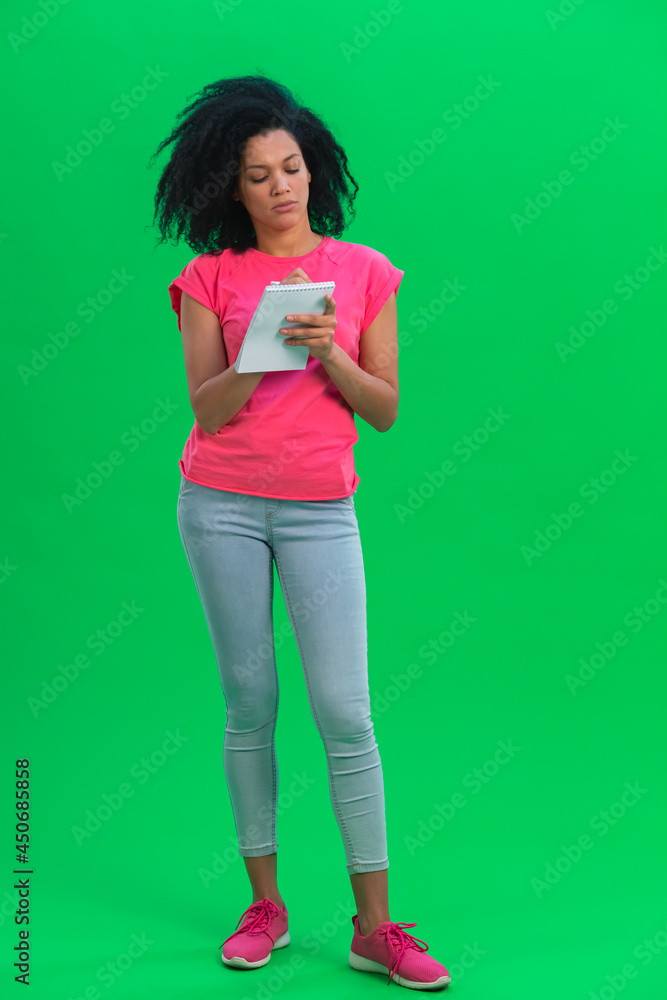 Portrait of young female African American thinking, then happy writing with pen in notebook. Black woman with curly hair poses on green screen in the studio. Full length. Portrait of young female African American thinking, then happy writing with pen in notebook. Black woman with curly hair poses on green screen in the studio. Full length.