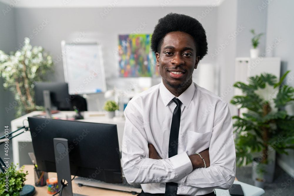Smiling friendly dark skinned afro men sits on desk wearing white shirt and tie, in background scenery of company, office boss, president, manager, businessman, arms crossed on stomach Smiling friendly dark skinned afro men sits on desk wearing white shirt and tie, in background scenery of company, office boss, president, manager, businessman, arms crossed on stomach