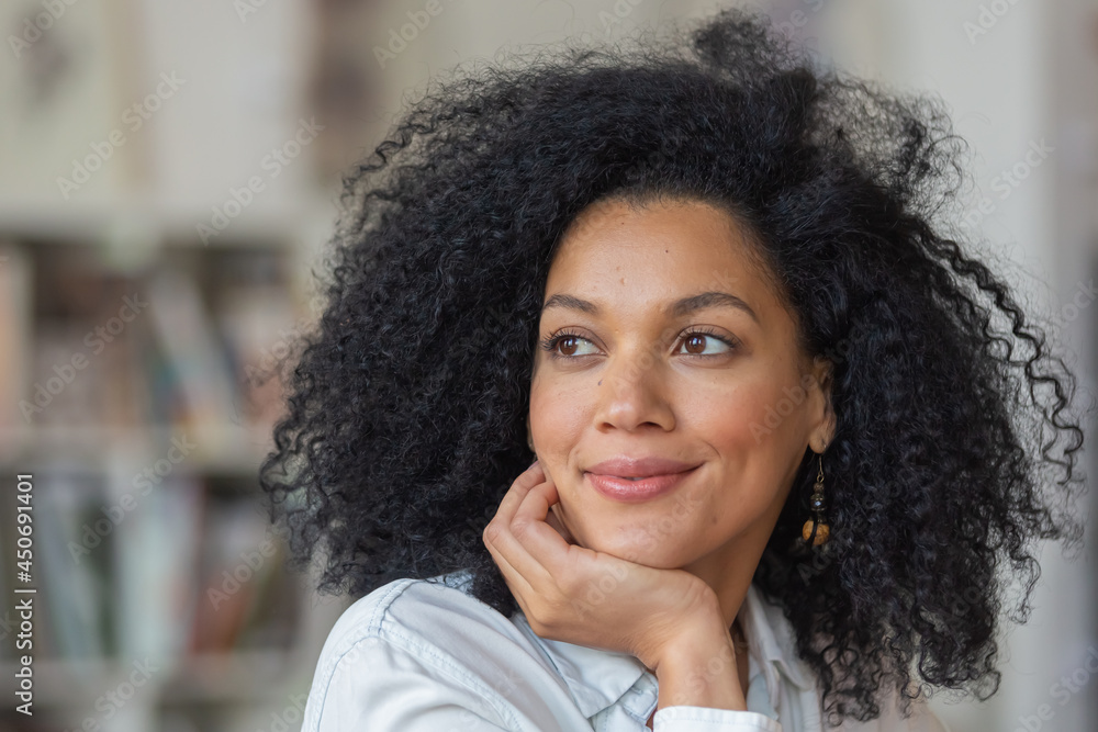 Portrait of a young African American woman looking to the side and smiling. Brunette with curly hair in white blouse sits at a table in light home office. Close up. Portrait of a young African American woman looking to the side and smiling. Brunette with curly hair in white blouse sits at a table in light home office. Close up.