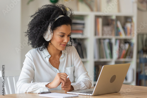 Portrait of African American woman talking on video conference call using laptop and headphones taking notes on notepad. Brunette sits at table in home office. Close up. photo