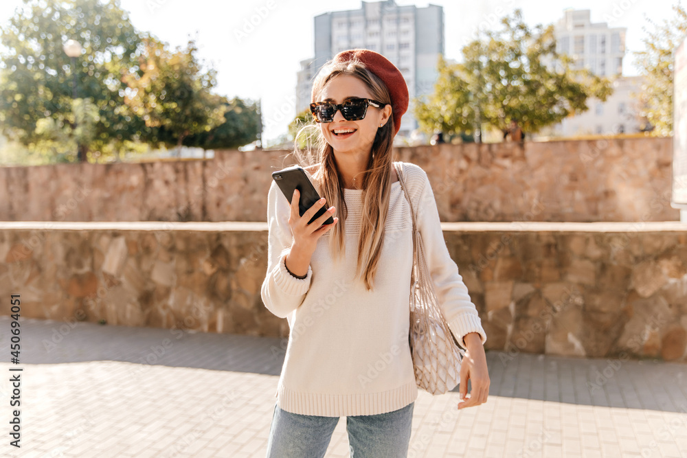Active french girl in sunglasses and beret chilling outdoor. Pleasant long-haired woman with phone walking around city.