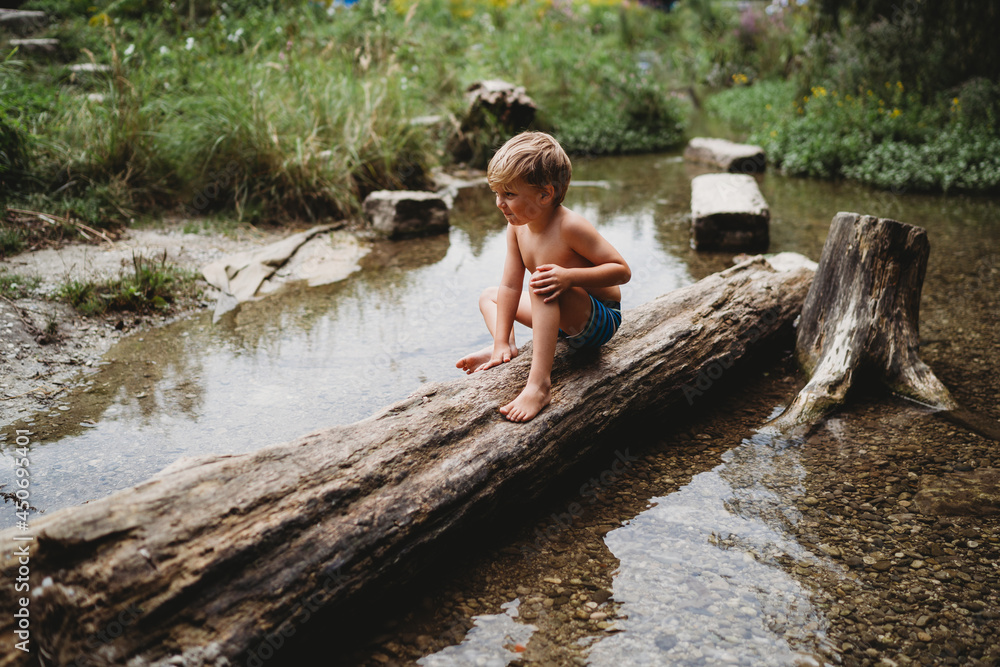 Young boy sitting on log in water in summer looking thoughtful Young boy sitting on log in water in summer looking thoughtful