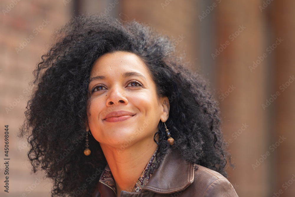 Portrait of stylish brooding young African American woman focused thinking about something. Brunette in brown leather jacket posing on street against backdrop of blurred brick building. Close up. Portrait of stylish brooding young African American woman focused thinking about something. Brunette in brown leather jacket posing on street against backdrop of blurred brick building. Close up.