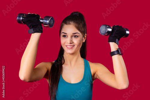 Young sporty girl smiling with long hair practicing fitness exercises raised hands up with two iron dumbbells.