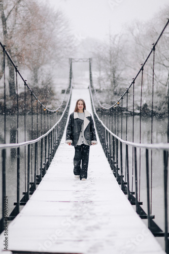A woman walks over the river on a suspension bridge in winter day. Young girl in warm clothes stands on a wooden footbridge in cold snowy day